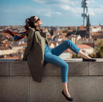 Woman reclining on a wall with city background on a sunny day. She wears sunglasses, an olive mac and blue skinny jeans. one leg is raised and she wears Angles barefoot ballet flats