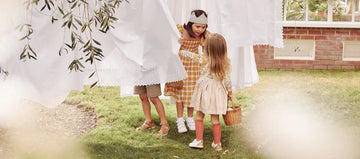 Three children in neutral summer clothes in a garden playing amongst a line of white laundry wearing Bobux barefoot shoes
