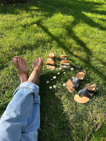Legs in jeans relaxes on sunny grass, daisies, tree shadows. Includes an array of barefoot sandals