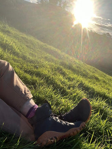 Person reclining on grass at sundown wearing Vivobarefoot Tracker boots from Happy Little Soles