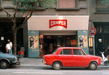 street in 1960s featuring red car and Camper storefront