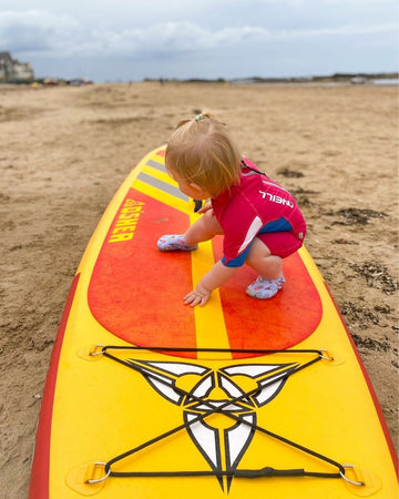 A small child wearing a rash vest crouches on a yellow and red paddleboard on a sandy beach, with cloudy skies and a distant coastline in the background.