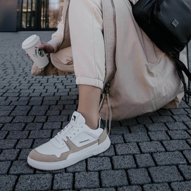 Person wearing Be Lenka Adults Barebarics Vegan Trainers Zing - White and Beige with beige joggers, coat and bag holding a coffee