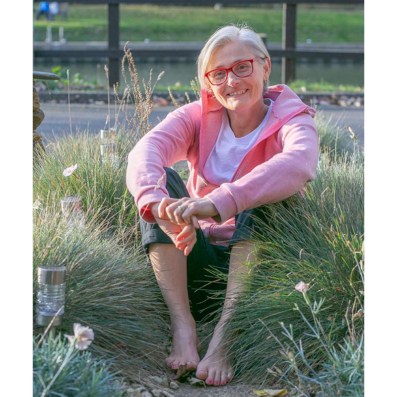 Bernie Landels sitting outdoors in a natural setting, barefoot, wearing a pink hoodie and glasses.